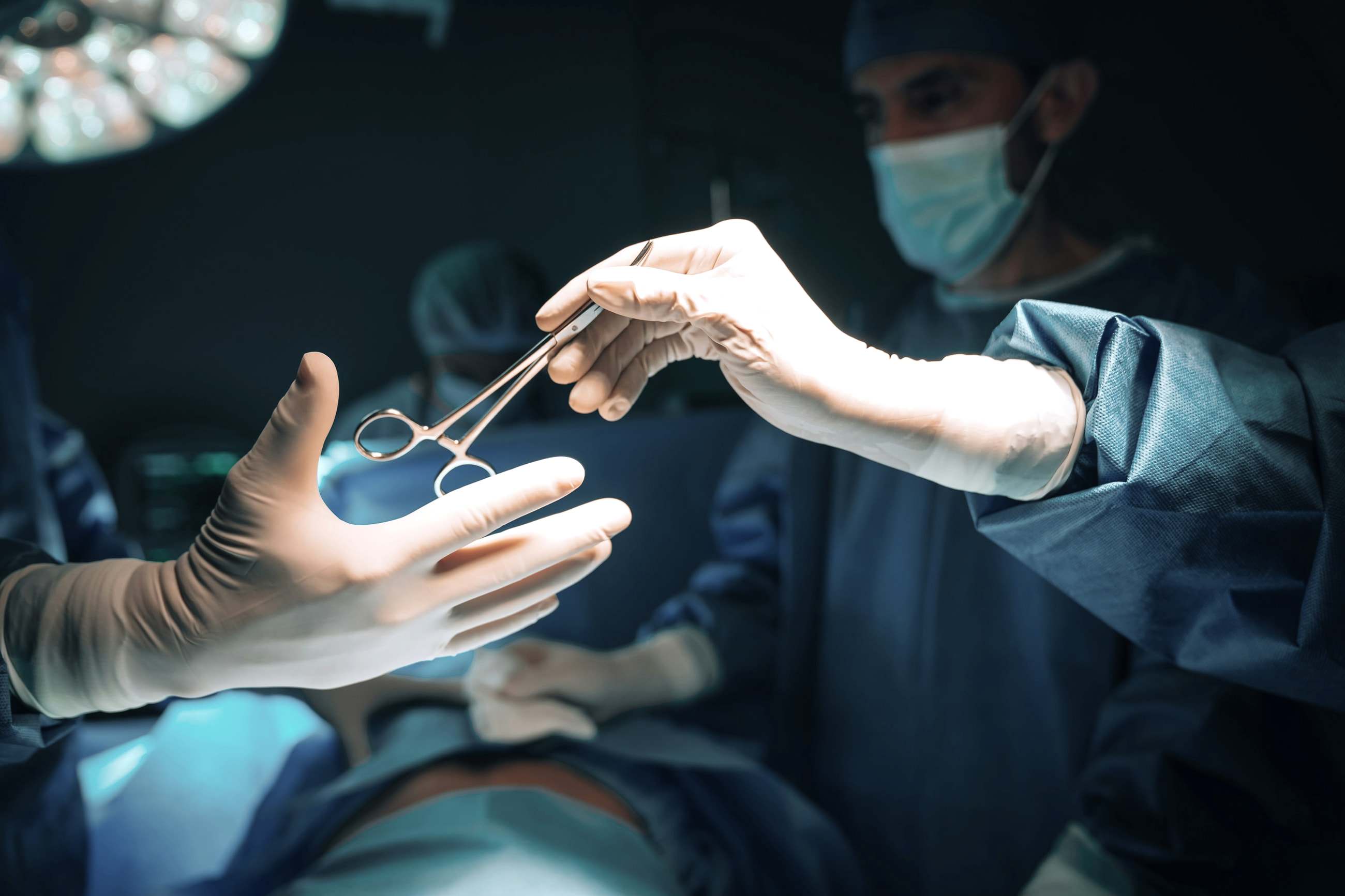 Mãos com luvas cirúrgicas passando uma pinça durante uma cirurgia.