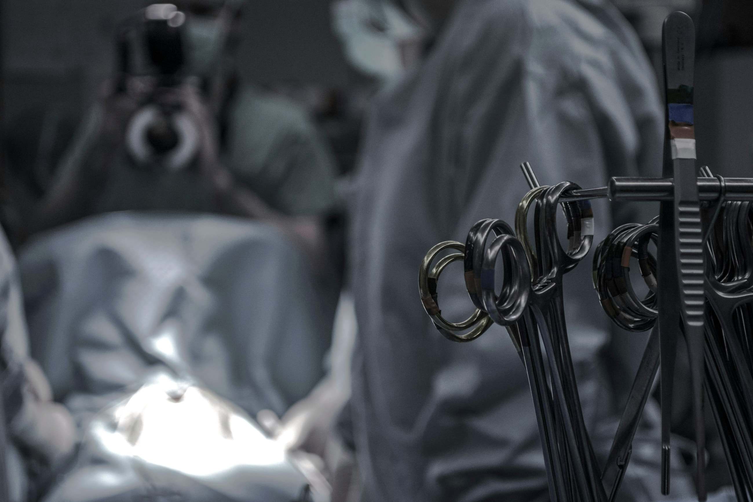 Pinças penduradas em suporte na sala de cirurgia, com a equipe cirúrgica desfocada ao fundo.