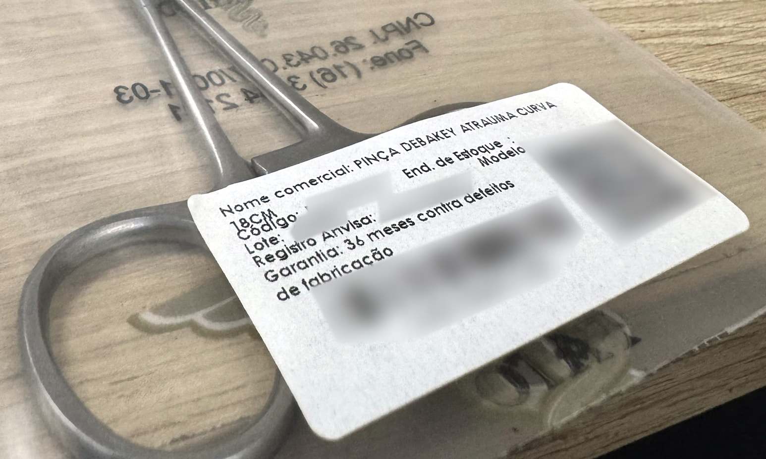 Etiqueta de uma pinça DeBakey embalada, com os campos nome comercial, código, lote, registro Anvisa e garantia de 36 meses; os dados de identificação, o código de barras e o QR code estão desfocados.